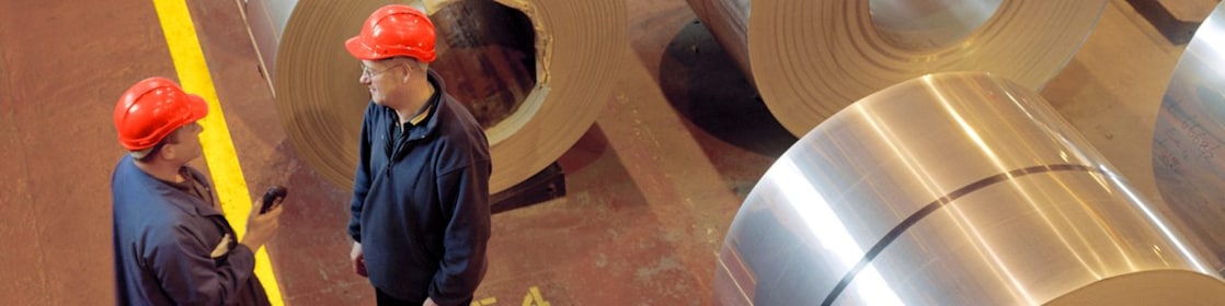 Two factory workers standing next to rolls of steel