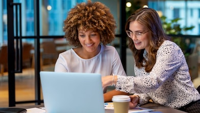 Two professionals collaborating at a table with a laptop, documents, and a coffee cup in a modern office setting, with large windows and contemporary furniture in the background.